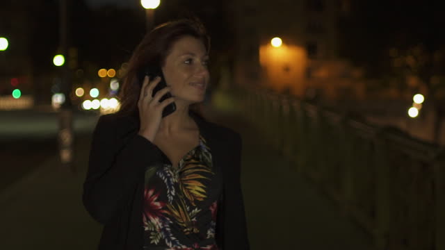 Glückliche-kaukasische-moderne-Frau-trägt-Blumenkleid,-schwarze-Jacke-und-rote-Haare-zu-Fuß-durch-die-Straße-und-telefonieren-auf-ihrem-Smartphone-in-der-Nacht.-Paris-4K-UHD.-Zeitlupe.