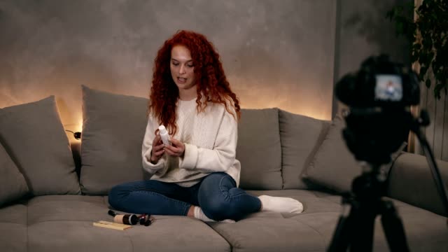 Curly-red-headed-young-girl-vlogger-is-talking-in-front-of-camera-recording-video-for-online-blog-in-internet,-demosrating-cosmetics.-Woman-is-wearing-jeans-and-white-sweater.-Sitting-in-a-living-room