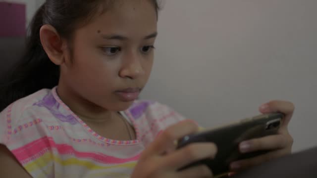 Adorable-chica-jugando-juegos-en-el-teléfono-inteligente-móvil-y-sentado-en-el-sofá-durante-la-cuarentena-en-casa.-Asiática-adolescente-enviando-mensajes-de-texto-sobre-comunicación-social-en-línea-con-el-teléfono-celular.
