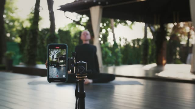 Bald-male-in-black-sportswear-is-performing-yoga-sitting-on-mat-at-landscaped-patio.-Shooting-himself-on-video-using-smartphone-on-tripod.-Close-up