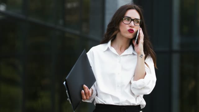Girl-in-glasses-and-white-shirt-tanding-near-business-center-with-papers-on-clipboard.-Smiling,-talking-on-cellphone-discussing-new-contract.-Close-up