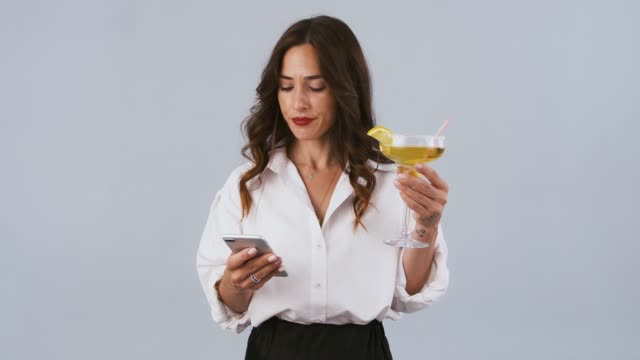 Lady-in-white-shirt-and-black-skirt-is-typing-on-her-smartphone-and-holding-cocktail-while-posing-on-gray-studio-background.-Close-up,-copy-space