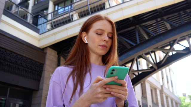 Joyful-pretty-redhead-girl-with-closed-eyes-listening-music-using-wireless-headphones-in-city-street