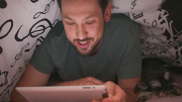 Close-up-of-face-of-a-young-bearded-and-mustashed-man,-using-digital-tablet-under-blanket,-who-is-chatting-and-waves-hand,-in-his-bed,-at-night.-His-yorkshire-terrier-is-lying-next-to-him