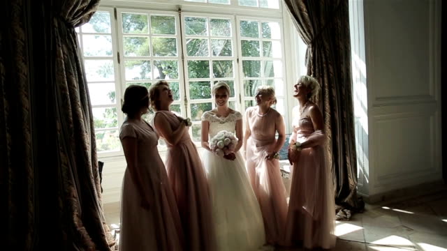 Bridesmaids-laughing-happy-near-bride-slow-motion.-Beautiful-girlfriends-pose-surrounding-bride-amusing-and-talking-in-sun-light-room.-Bachelorette-party-emotional.-Elegant-wedding-dress-fashion-style