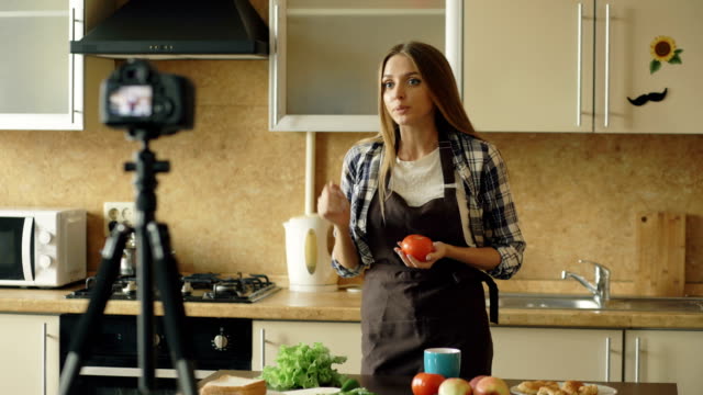 Young-attractive-woman-in-apron-shooting-video-food-blog-about-cooking-on-dslr-camera-in-kitchen
