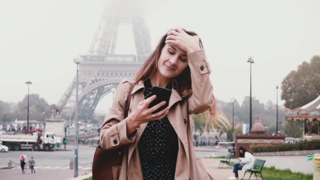 Young-beautiful-woman-walking-near-the-Eiffel-tower-in-Paris,-France-and-using-the-smartphone,-browsing-the-Internet