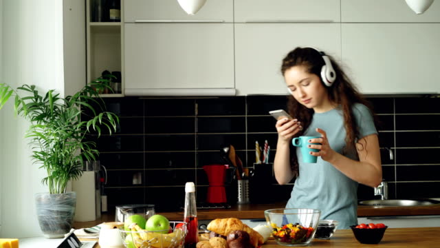 Attraktive-glückliches-Mädchen-tanzen-und-singen-in-Küche-während-mit-Smartphone-und-Musikhören-zu-Hause-am-Morgen
