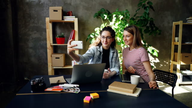 Jóvenes-empresarias-están-haciendo-selfie-juntos-en-la-oficina-moderna-mientras-está-sentado-en-el-escritorio.-Que-estén-posando-con-caras-divertidas-y-sonrientes,-luego-viendo-fotos.