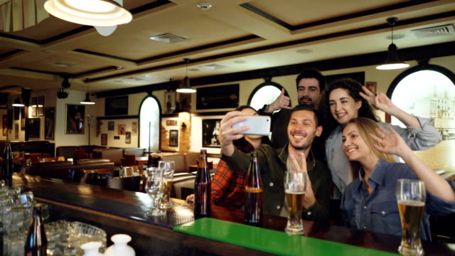 Amigos-están-tomando-selfie-con-smartphone-en-la-barra.-Jóvenes-plantean,-riendo-y-hablando.-Botellas-de-cerveza-y-vasos-en-primer-plano