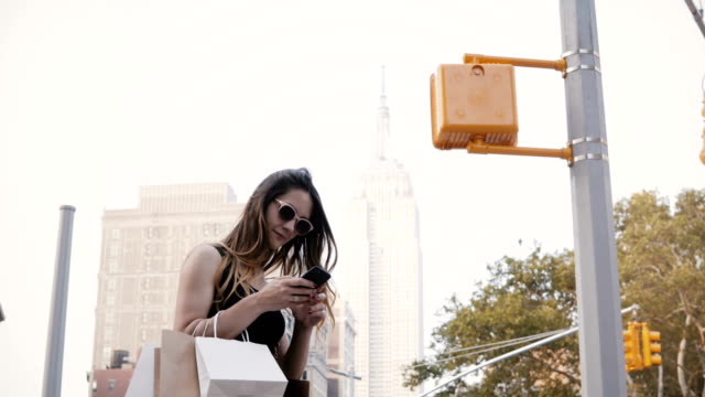 Happy-beautiful-Caucasian-businesswoman-with-shopping-bags-in-sunglasses-using-smartphone-app-near-Empire-State-Building