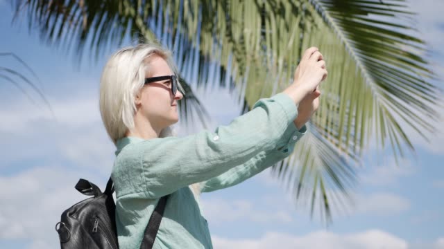 Young-beautiful-slim-woman-with-long-blonde-hair-in-sunglasses-and-green-shirt-standing-near-palm-tree-and-take-a-pictures-by-smartphone-on-a-blue-sky-background.-Girl-using-mobile-phone