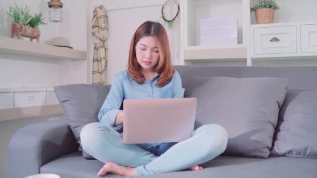 Porträt-der-schönen-attraktiven-jungen-lächelnden-Asiatin-mit-Computer-oder-Laptop-auf-dem-Sofa-liegend-beim-Entspannen-im-Wohnzimmer-zu-Hause.-Genießen-Zeit-Lifestyle-Frauen-bei-Wohnkonzept.