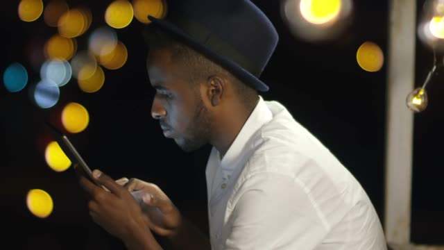 Chico-africano-alegre-uso-de-tableta-en-azotea-en-la-ciudad-de-noche