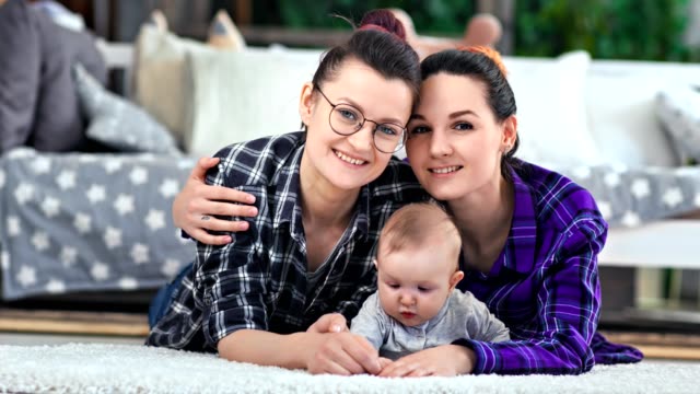 Two-beautiful-hipster-female-parent-posing-with-little-cute-baby-lying-on-floor-looking-at-camera