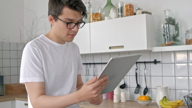 Attractive-man-at-home-using-tablet-in-kitchen-sending-message-on-social-media-smiling-enjoying-modern-lifestyle-wearing-white-shirt