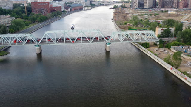 Vista-aérea-del-puente-ferroviario,-con-un-tren-en-movimiento-en-él,-a-través-del-río-que-fluye-a-través-de-una-ciudad-importante