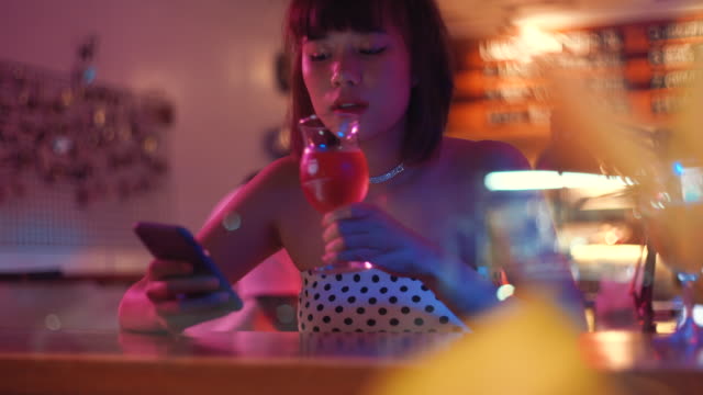 Young-asian-millennial-woman-using-smartphone-and-drinking-beer-in-nightclub-bar,-lifestyle-concept