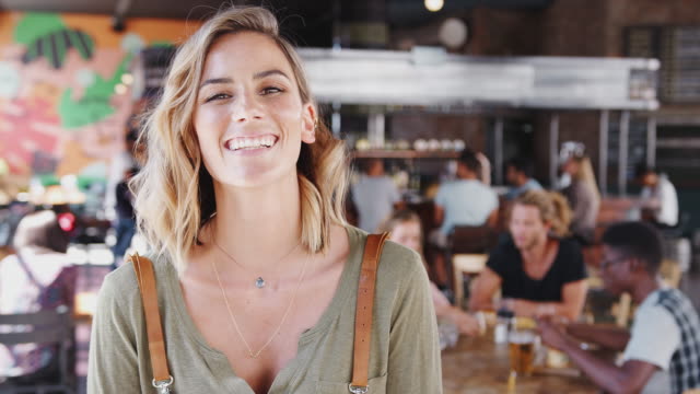 Portrait-of-waitress-wearing-apron-holding-digital-camera-in-busy-bar-restaurant-smiling-at-camera---shot-in-slow-motion