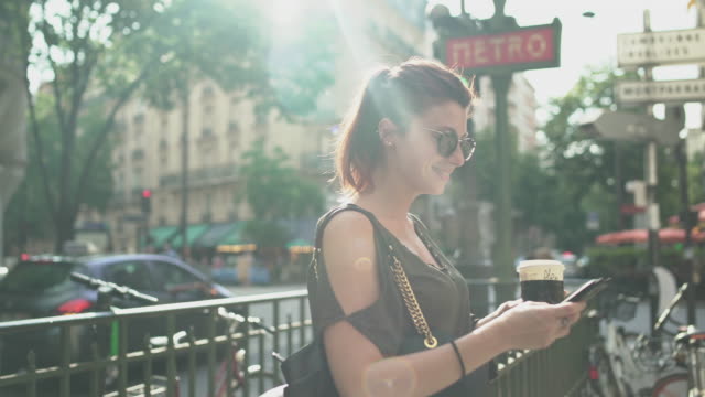 Young-attractive-smiling-woman-writing-a-text-message-on-her-smartphone-at-the-subway-exit-in-street,-holding-her-coffee,-during-sunny-summer-in-Paris.-Freckles,-sunglasses,-piercings,-red-hair,-4K.