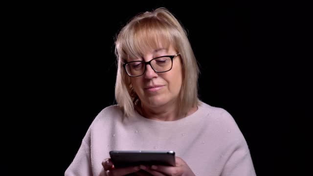 Closeup-shoot-of-old-caucasian-blonde-female-in-glasses-using-the-tablet-in-front-of-the-camera-with-background-isolated-on-black