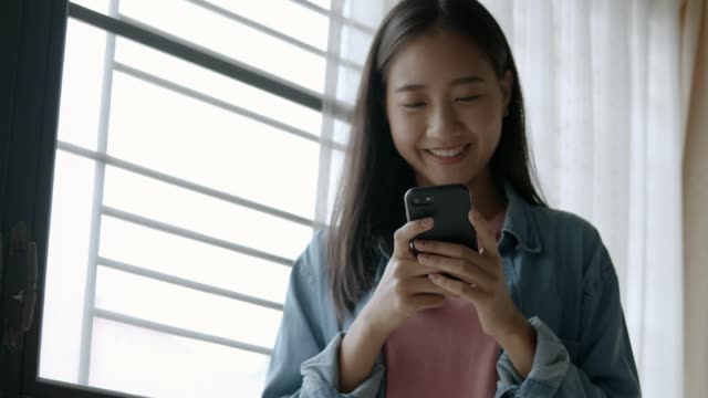 Portrait-smiling-young-asian-business-woman-holding-typing-mobile-phone-and-scrolls-through-social-media-feed-in-smartphone-standing-beside-window-at-home-office.