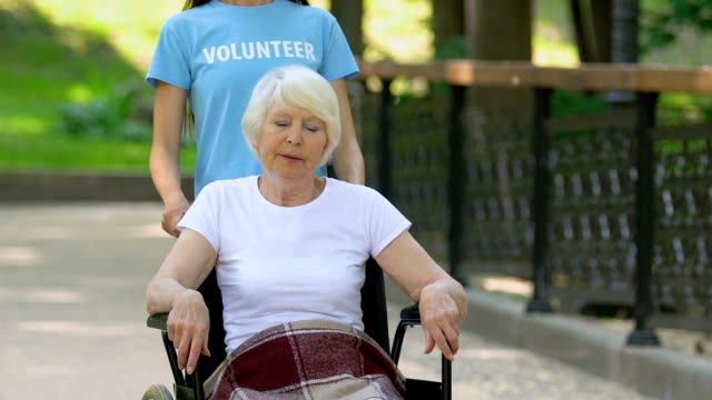 Volunteer-helping-elderly-female-patient-in-wheelchair,-walking-in-hospital-park