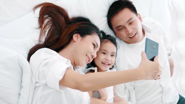 Asiatische-Familie-lacht-und-schaut-in-Smartphone,-während-auf-dem-Bett-im-Schlafzimmer-liegen