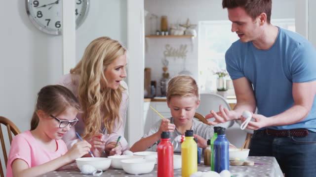 Familie-sitzt-um-Küchentisch-mit-Eltern,-die-Kindern-helfen,-Eier-für-Ostern-zu-malen---in-Zeitlupe-gedreht
