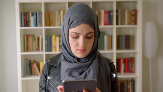 Nahaufnahme-Porträt-von-jungen-attraktiven-muslimischen-Studentin-in-Hijab-mit-dem-Tablet-Blick-auf-die-Kamera-in-der-College-Bibliothek-in-Innenräumen