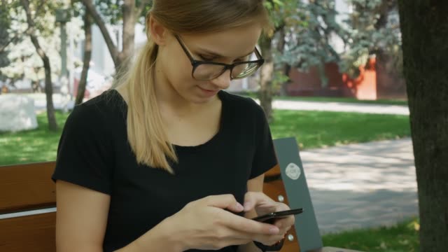 Pretty-caucasian-woman-in-eyeglasses-use-of-black-mobile-phone-and-sit-on-the-bench-in-the-city-park