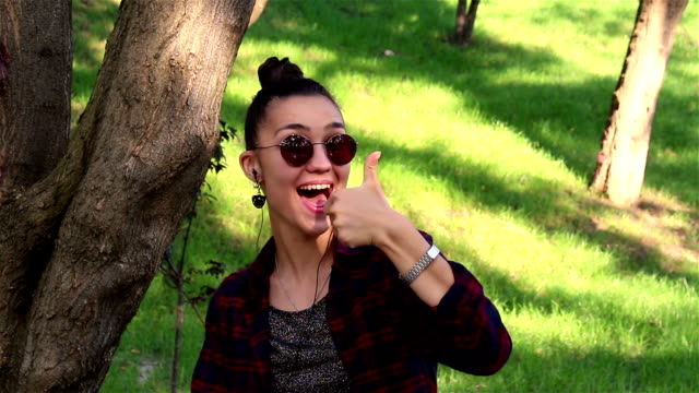 Beautiful-girl-in-sunglasses,-actively-laughing,-and-showing-thumb-up-while-sitting-on-a-bench-in-the-park