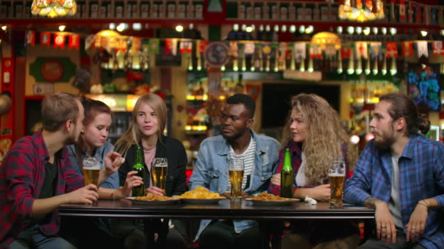 Un-grupo-multiétnico-de-hombres-y-mujeres-jóvenes-beben-cerveza-en-un-bar-y-comen-patatas-fritas-y-debaten-alegremente-sobre-la-universidad.-Reírse-de-una-broma