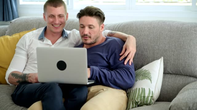 Pareja-gay-relajándose-en-el-sofá-usando-la-computadora-portátil.-Mecanografía.