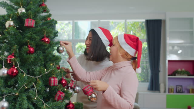 Asiatische-Freundinnen-schmücken-Weihnachtsbaum-auf-Demonstaus.-Weibliche-teen-glücklich-lächeln-feiern-Weihnachts-Winterurlaub-zusammen-im-Wohnzimmer-zu-Hause.