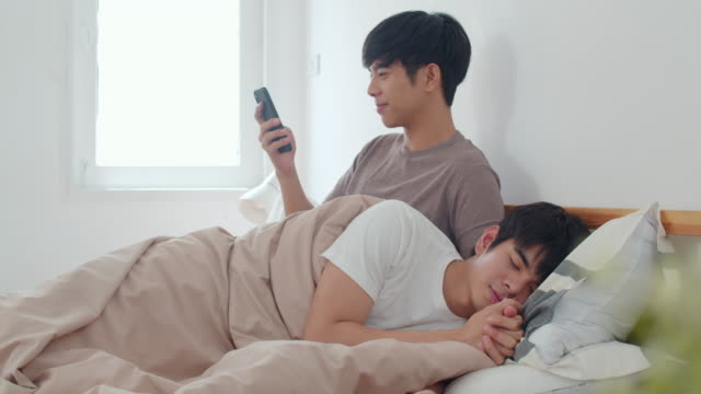 Pareja-gay-asiática-usando-el-teléfono-móvil-en-casa.-Joven-Asia-LGBTQ+-hombre-feliz-descanso-después-de-despertar,-comprobar-las-redes-sociales-mientras-su-novio-duerme-acostado-en-la-cama-en-el-dormitorio-en-casa-en-el-concepto-de-la-mañana.