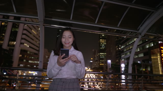 Joven-mujer-asiática-sonriente-de-negocios-usando-teléfono-inteligente-en-el-centro-de-la-ciudad-moderna-en-la-noche-de-Bangkok-Tailandia.-Comunicación-Concept-Technology-por-teléfono-móvil