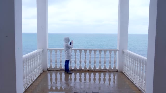 Mujer-en-la-chaqueta-blanca-toma-video-de-las-olas-del-mar-en-el-teléfono-inteligente-en-la-hermosa-terraza-con-vista-al-mar.-Vista-trasera.