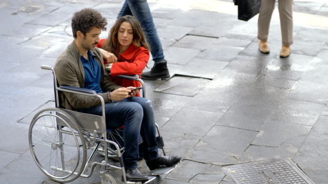 Woman-kneeling-near-his-boyfriend-on-wheelchair-looking-on-smartphone-outdoor