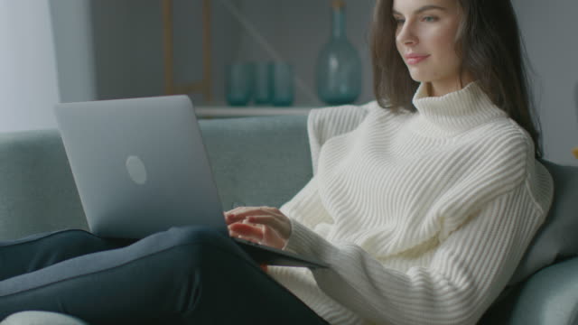 Beautiful-Young-Woman-Works-on-Laptop-Computer-while-Sitting-on-the-Chair.-Sensual-Girl-Wearing-Sweater-Works-On-Notebook;-Studies,-Surfs-Internet,-Uses-Social-Media-while-Relaxing-in-Cozy-Apartment