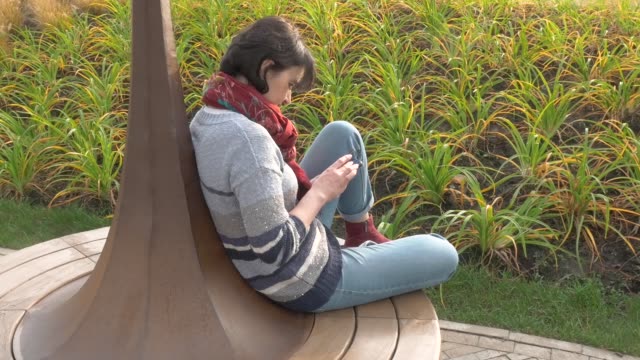 Caucasian-woman-sits-on-a-round-bench,-clicks-with-a-phone-in-her-hands