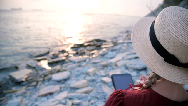 Close-up-View-from-the-back-An-attractive-young-girl-in-a-summer-red-dress-and-a-straw-hat-sits-on-a-stone-by-the-sea-at-sunset-and-watches-something-on-a-tablet.-Swipe-across-the-screen