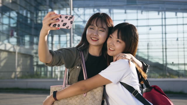 Lächelnd-elated-junge-asiatische-Frauen-in-modernen-Kleidern-umarmen-und-selfie-in-der-Nähe-des-Flughafens