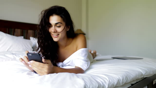 Junge-Frau-mit-Zelle-Smartphone-glücklich-lächelnde-schöne-Mädchen-Schlafzimmer-morgens-am-Bett-liegen