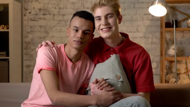 Two-multi-ethnic-homosexual-friends-sit-on-the-couch-holding-hands,-hugging,-smiling,-looking-at-the-camera.-Home-cosiness,-family,-lgbt.-60-fps