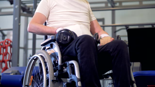 A-disabled-man-is-sitting-in-a-wheelchair-with-dumb-bells,-moving-his-shoulders-and-talking