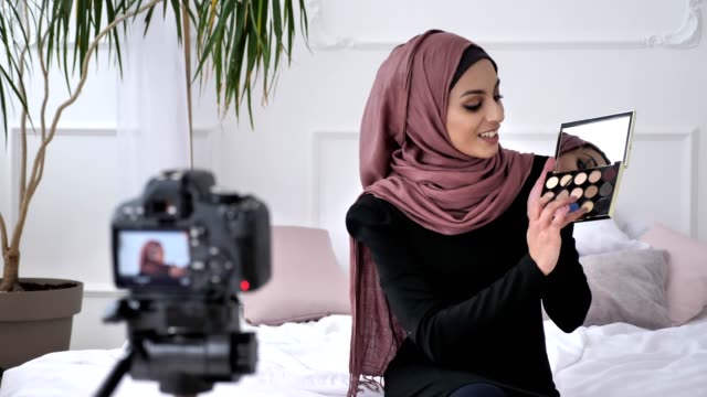 Joven-hermosa-chica-India-en-blogger-hijab-hablando-en-la-cámara,-sonriendo,-mostrando-una-nueva-compra,-cosméticos,-sombra-de-ojos-paleta,-home-comfort-en-el-fondo.-60-fps