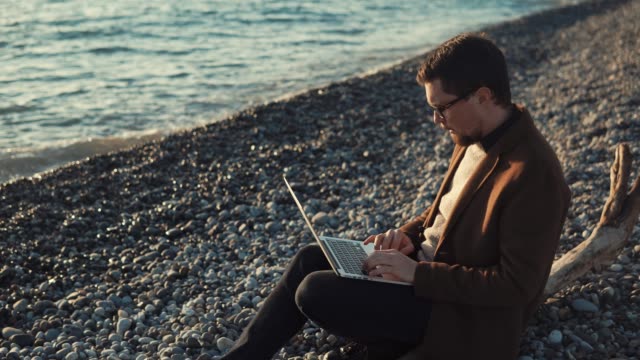 Hombre-con-barba-está-escribiendo-texto-en-un-ordenador-portátil,-sentado-en-una-orilla-del-mar,-vista-lateral