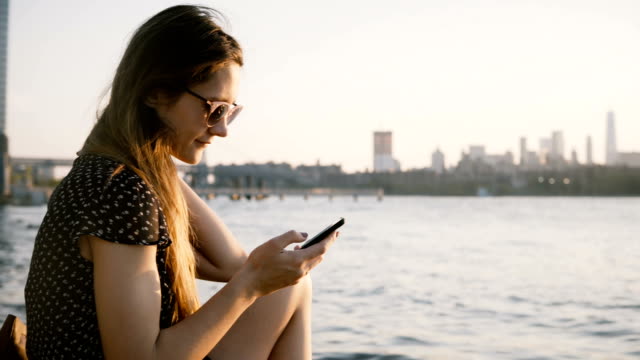 Nachdenklich-Kaukasische-Mädchen-in-Sonnenbrille-mit-Smartphone-app-für-soziale-Netzwerke-erstaunlichen-Sonnenuntergang-am-Stadtstrand-4K