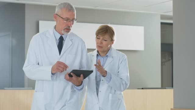In-the-Hospital-Doctors-have-Discussion-while-Using-Tablet-Computer.-They're-Walking-through-the-Hallway.-In-the-Background-Patients-and-Medical-Personnel.-New-Modern-Fully-Functional-Medical-Facility.